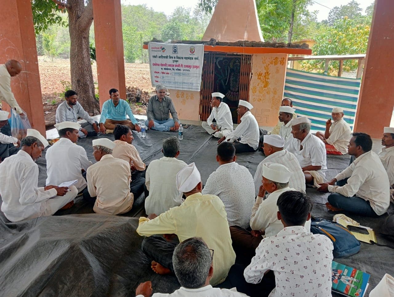 गावातील मंदिराच्या ओसरीवर ताडपत्रीवर मांडी घालून बसलेले सुमारे तीस गावकरी अधिकाऱ्यांचे मार्गदर्शन ऐकताना; मागील फलकावर 'शबरी आदिवासी वित्त व विकास महामंडळ मर्या., पुणे'