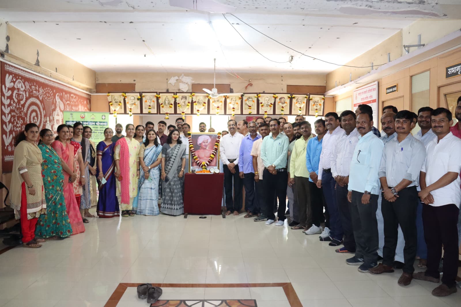 जागतिक आदिवासी दिनानिमित्त कार्यालयात बिरसा मुंडा यांच्या पुष्पहार घातलेल्या प्रतिमेभोवती उभे सुमारे पंचेचाळीस कर्मचारी; मागील भिंतीवर राष्ट्रपुरुषांच्या हारांकित प्रतिमा आणि डावीकडे वारली चित्रकला