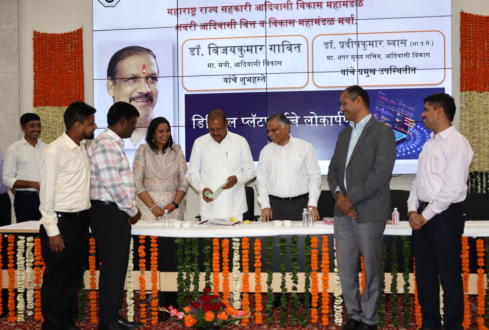 लोकार्पण सोहळ्यात सुमारे अकरा जण — व्यासपीठावर हिरवा दस्तऐवज हाताळणारे मान्यवर आणि दोन्ही बाजूंस उभे अधिकारी; मागील पडद्यावर कार्यक्रमाचा संपूर्ण तपशील