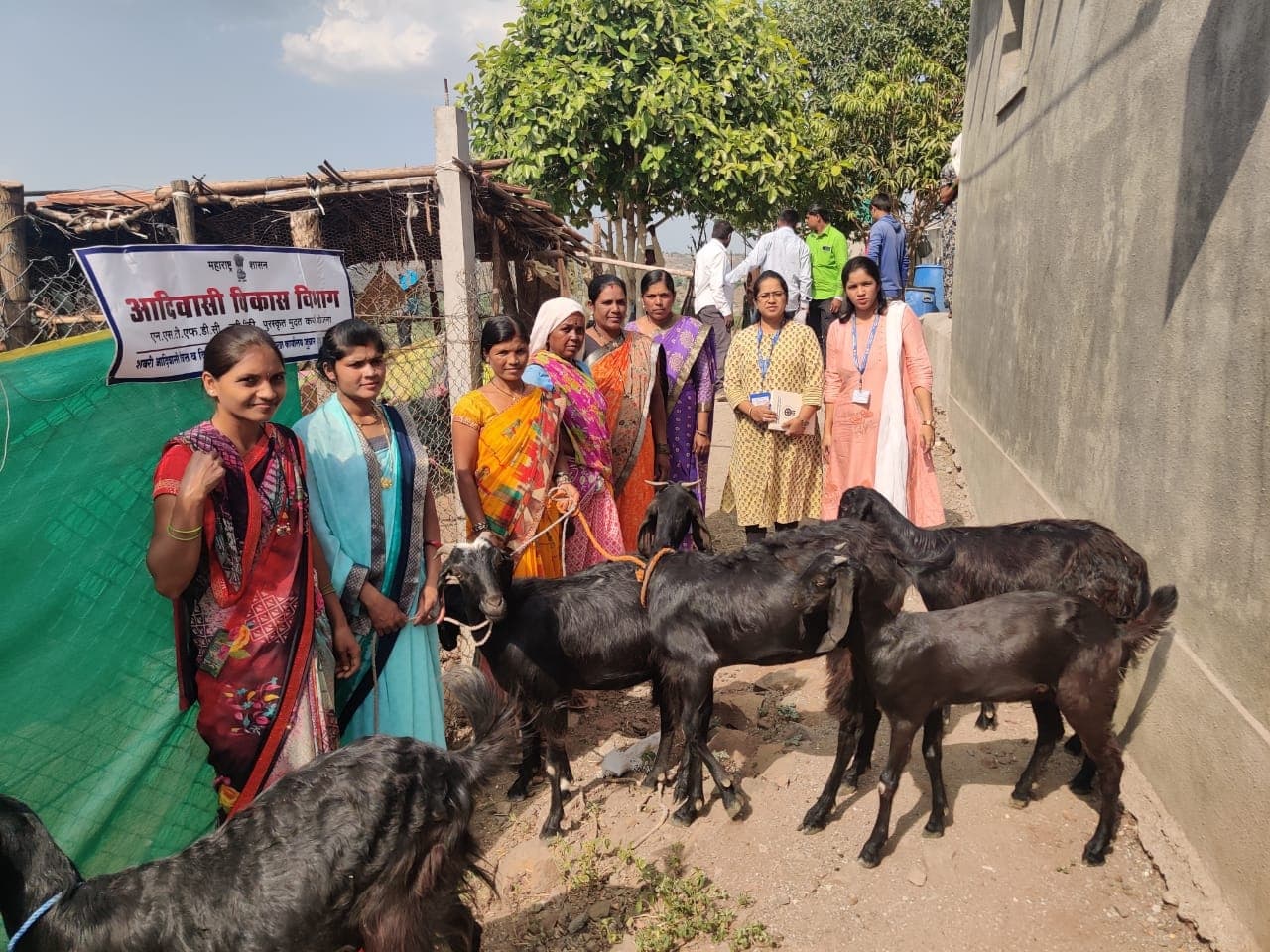 गावातील घराशेजारी शेळ्यांसह उभ्या असलेल्या आठ महिला व दोन क्षेत्रीय अधिकारी; कुंपणावरील फलकावर 'एन.एस.टी.एफ.डी.सी. पुरस्कृत मुदत कर्ज योजना — शबरी आदिवासी वित्त व विकास महामंडळ'