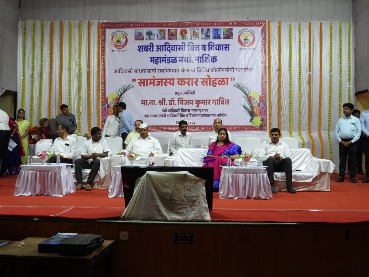 सामंजस्य करार सोहळ्यासाठी सजवलेले व्यासपीठ — लाल गालिच्यावर पांढऱ्या खुर्च्यांवर बसलेले सुमारे नऊ मान्यवर आणि मागील फलकावर मंत्री डॉ. विजयकुमार गावित यांच्या प्रमुख उपस्थितीचा उल्लेख