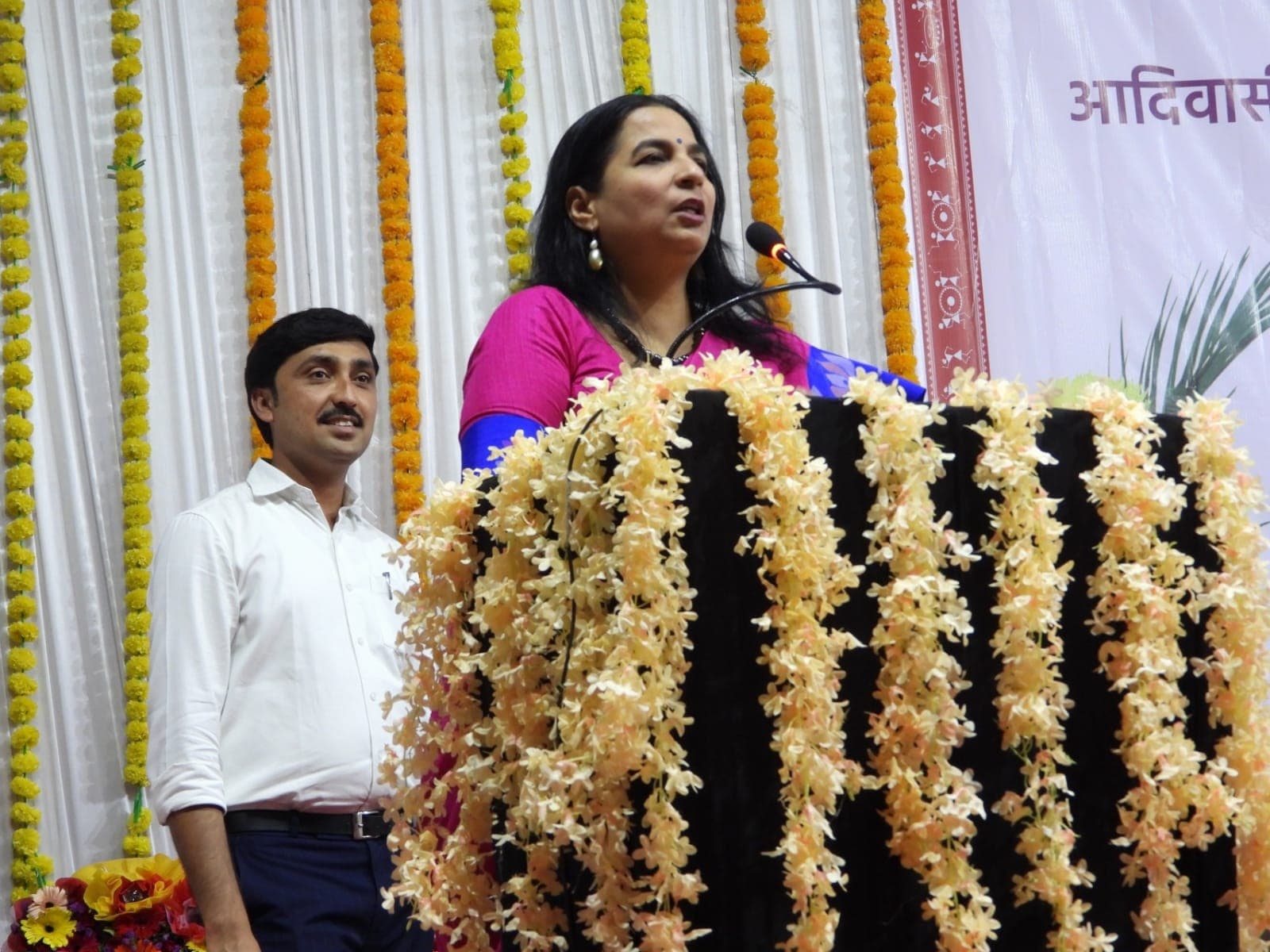 फुलांच्या माळांनी सजवलेल्या व्यासपीठावरून ध्वनिवर्धकात बोलणाऱ्या महिला अधिकारी; शेजारी ऐकत उभा असलेला सहकारी आणि मागे वारली नक्षीचा पडदा