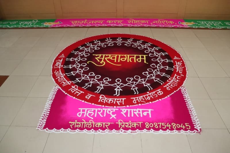 जमिनीवरील रांगोळी — लाल-मरून वर्तुळाभोवती हातात हात घालून उभ्या वारली शैलीतील पांढऱ्या आकृत्या, मध्यभागी 'सुस्वागतम' आणि कडेने महामंडळाचे नाव