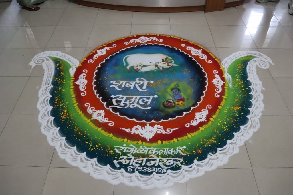 जमिनीवर काढलेली मोठी गोलाकार रांगोळी — निळ्या मध्यभागी पांढरी गाय व वासरू आणि मडके घेतलेला कृष्ण, मध्यभागी 'शबरी सुमुल' अशी अक्षरे