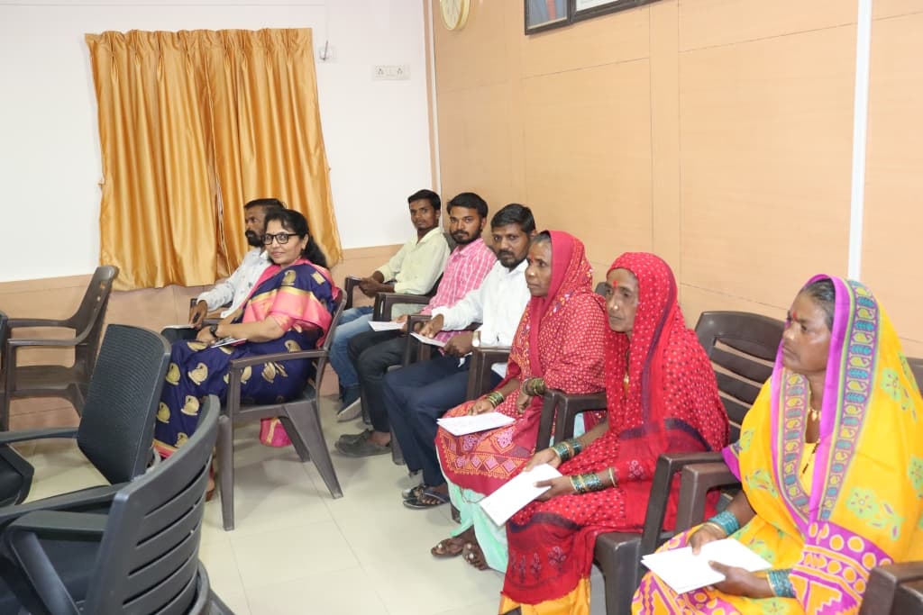 बैठकीच्या खोलीत खुर्च्यांवर बसलेले सुमारे नऊ जण — त्यात पारंपरिक साड्या व डोक्यावर पदर घेतलेल्या चार आदिवासी महिला, बहुतेकांच्या हातात कागद