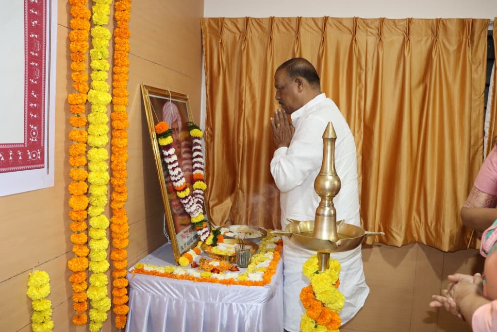 पगडी घातलेल्या मान्यवरांच्या हारांकित प्रतिमेसमोर हात जोडून उभे पांढऱ्या कुर्त्यातील गृहस्थ; शेजारी नुकतीच प्रज्वलित केलेली समई