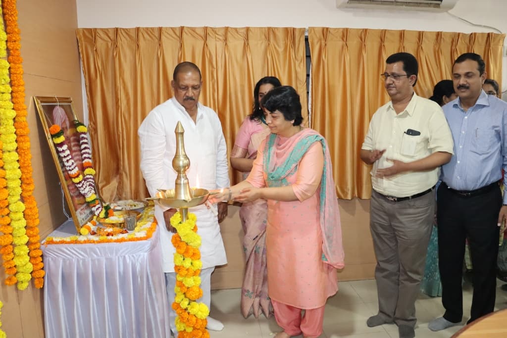 उभी समई तेवत्या वातीने प्रज्वलित करणाऱ्या महिला अधिकारी; शेजारी पांढऱ्या कुर्त्यातील मान्यवर आणि डावीकडे हार घातलेली प्रतिमा