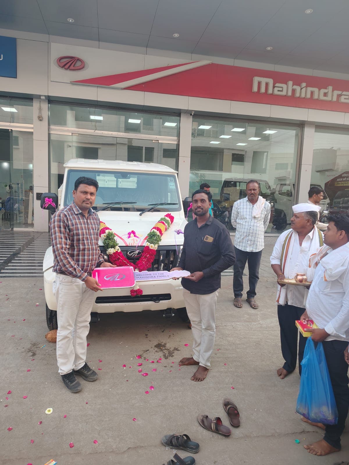 वाहन विक्री दालनात हार घातलेल्या पांढऱ्या बोलेरो पिकअपसमोर मोठे गुलाबी चावी-कार्ड धरून उभे दोन जण; उजवीकडे पूजेचे ताट घेतलेले पुरोहित व मिठाईचा पुडा