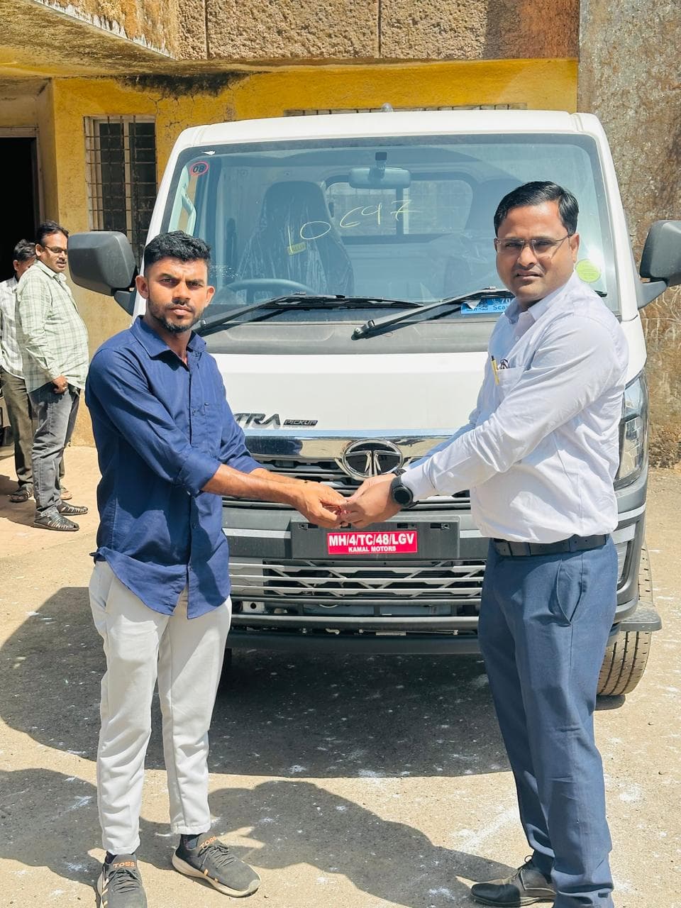 शासकीय इमारतीसमोर नव्या पांढऱ्या टाटा इंट्रा पिकअपच्या बोनेटवर हस्तांदोलन करत चावी सुपूर्द करणारे दोन जण