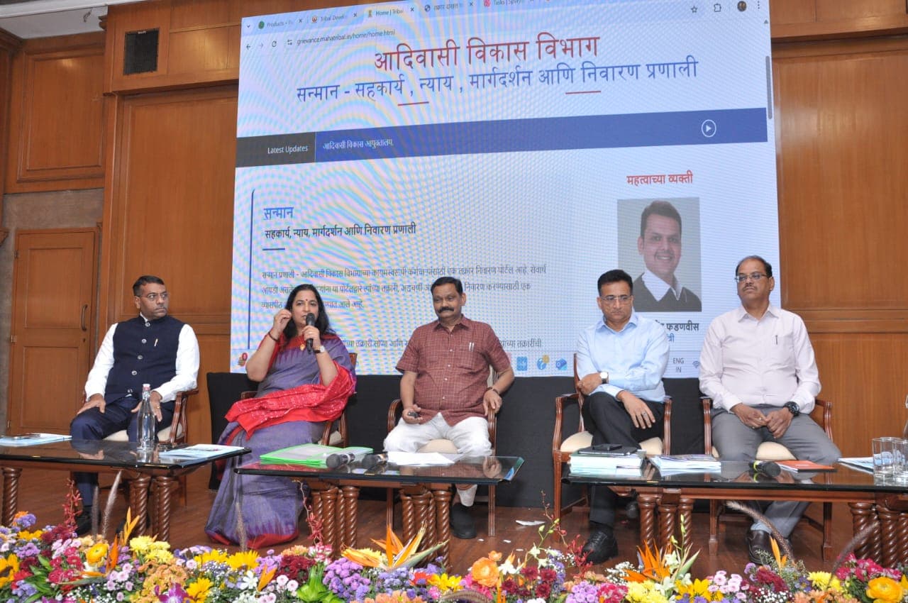 लाकडी सभागृहात फुलांनी सजवलेल्या व्यासपीठावर बसलेले पाच अधिकारी आणि ध्वनिवर्धकात बोलणाऱ्या जांभळ्या साडीतील महिला अधिकारी; मागील पडद्यावर विभागाचे 'सन्मान' तक्रार निवारण संकेतस्थळ