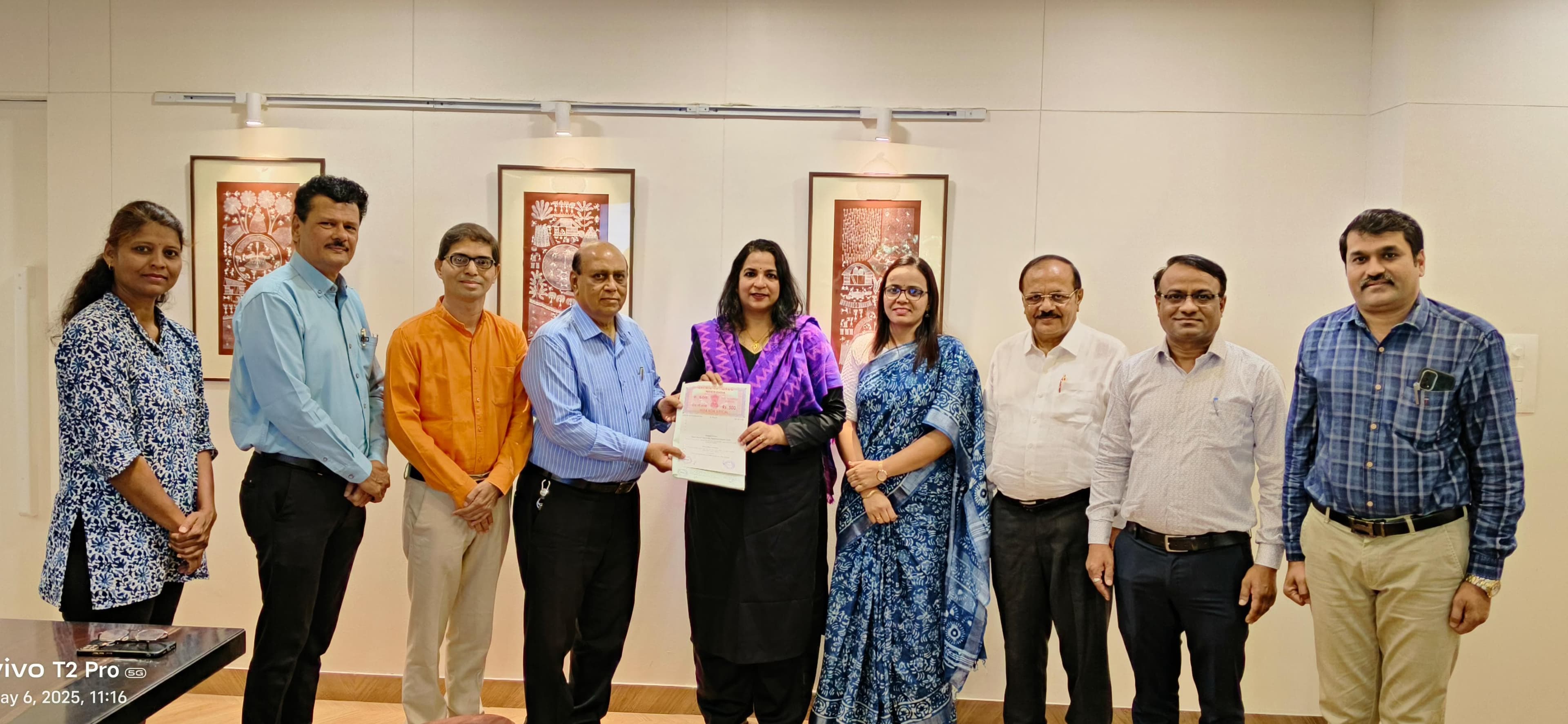तीन वारली चित्रे लावलेल्या कार्यालयात मुद्रांक कागदावरील करारपत्र सुपूर्द करताना उभे नऊ जण